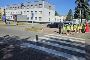 Zdjęcie przedstawia policjantkę oraz dzieci w kamizelkach odblaskowych przed przejściem dla pieszych.