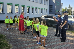 Zdjęcie przedstawia policjantki, nauczycielkę oraz dzieci w kamizelkach odblaskowych stojących na chodniku.