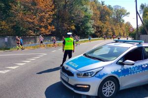 Zdjęcie przedstawia policjanta, radiowóz oraz uczestników biegu.