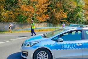 Zdjęcie przedstawia policjanta, radiowóz oraz uczestników biegu.