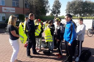 Zdjęcie przedstawia policjanta rozdającego kamizelki odblaskowe grupie rowerzystów.