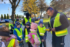 Zdjęcie przedstawia policjanta oraz dzieci w kamizelkach odblaskowych.