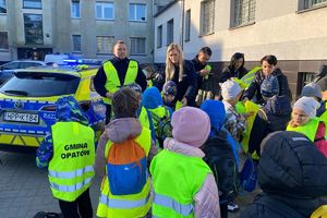 Zdjęcie przedstawia policjantów, dzieci w kamizelkach odblaskowych oraz radiowozy.