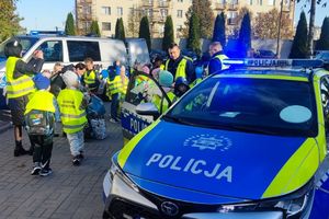 Zdjęcie przedstawia policjantów, dzieci w kamizelkach odblaskowych oraz radiowozy.