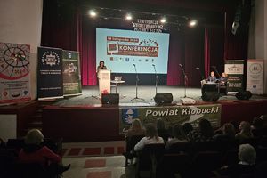 Zdjęcie przedstawia kobietę na scenie przy mównicy oraz siedzących na widowni uczestników konferencji.