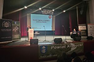 Zdjęcie przedstawia mężczyznę na scenie przy mównicy oraz siedzących na widowni uczestników konferencji.