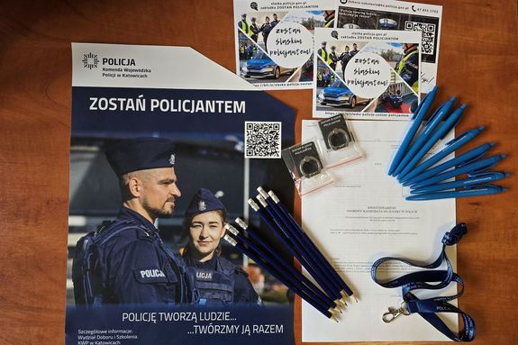 Zdjęcie przedstawia plakat oraz ulotki promujące wstąpienie do służby w policji, a także długopisy, ołówki i smycz.