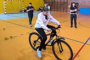 Zdjęcie przedstawia policjanta, nauczyciela oraz dziewczynę na rowerze w sali gimnastycznej.
