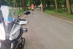 Zdjęcie przedstawia policyjny motocykl oraz uczestników biegu.