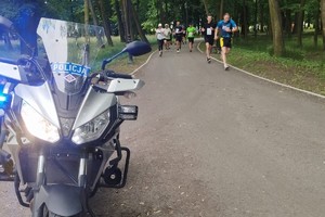 Zdjęcie przedstawia policyjny motocykl oraz uczestników biegu.