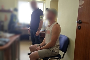 Zdjęcie przedstawia policjanta w ubraniu cywilnym, stojącego obok zatrzymanego, który siedzi na krześle i na ręce ma założone kajdanki.