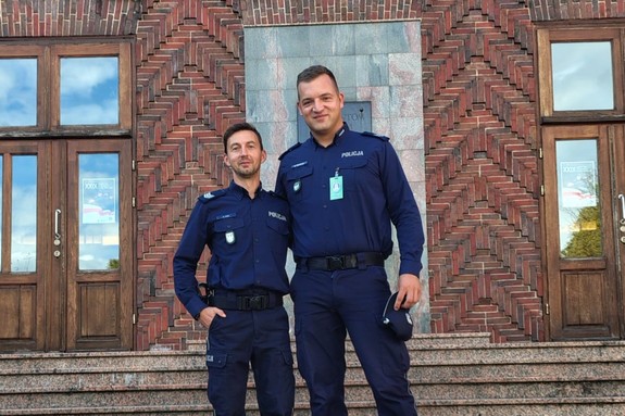 dwóch umundurowanych policjantów stoi przed budynkiem