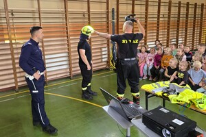 umundurowani  strażacy i policjant stoją przed dziećmi, prowadzą prelekcje, pokazują hełm strażacki