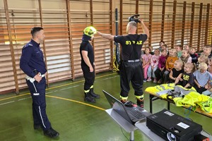 umundurowani  strażacy i policjant stoją przed dziećmi, prowadzą prelekcje, pokazują hełm strażacki