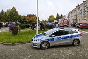Na zdjęciu radiowóz policyjny zaparkowany na parkingu przed budynkami mieszkalnymi. W tle widoczni są strażacy i pojazdy ratownicze.