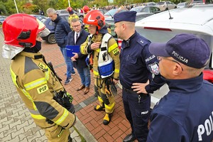 Na zdjęciu policjanci rozmawiają ze strażakami ubranymi w specjalistyczne kombinezony. Spotkanie odbywa się na parkingu przy budynkach mieszkalnych.