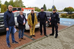 Na zdjęciu grupa osób, w tym policjanci i strażacy, stojąca przed budynkiem urzędu. W tle widać zaparkowane samochody.