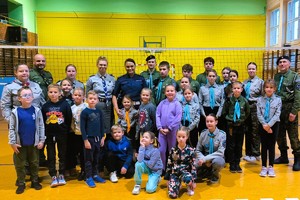 Grupa harcerzy i zuchów i policjantka pozuje wspólnie do zdjęcia w sali gimnastycznej po zakończonych zawodach sportowych.