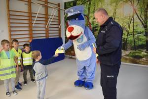 Policjant i maskotka Sznupek przekazują dzieciom kamizelki odblaskowe w sali gimnastycznej.
