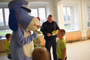 Policjant i maskotka Sznupek przekazują dzieciom kamizelki odblaskowe w sali gimnastycznej.