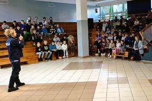 Policjantka prowadzi spotkanie z uczniami siedzącymi na trybunach sali gimnastycznej.