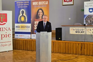 Mówca przy mównicy przemawia podczas konferencji na tle materiałów kampanii.