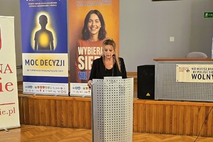 Kobieta przemawia przy mównicy, w tle banery kampanii.