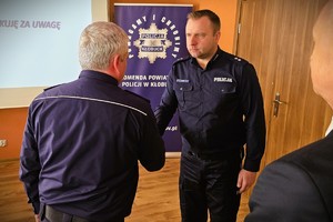 Odprawa służbowa policjantów, na zdjęciu widać grupę umundurowanych policjantów oraz kobiety i mężczyzn ubranych po cywilnemu, w tle rzutnik, na którym prezentowane są treści dotyczące bezpieczeństwa powiatu kłobuckiego