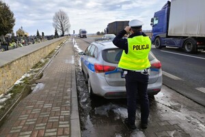 policjant w mundurze stoi przy radiowozie, obserwuje ciężarówki jadące ulicą